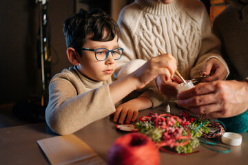 Close-up of pretty focused child boy writing number on red bag by brush and white paint sitting at table with family, making Christmas advent calendar together on xmas Eve, waiting for winter holiday.