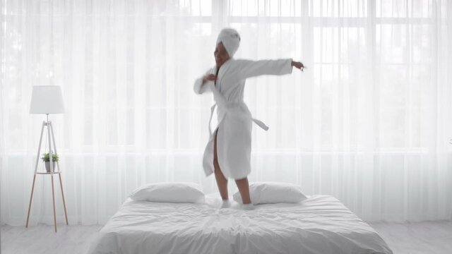 Happy Young African American Woman Wearing Bathrobe Jumping On Bed At Home