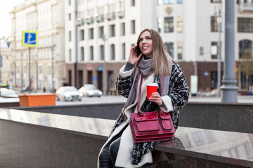 A young woman on a city street speaks on the phone with a glass of coffee in her hand. Beautiful blonde in a stylish warm coat. Walking in the cold season.