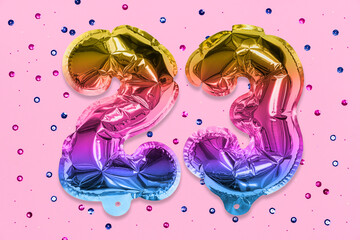 Rainbow foil balloon number, digit twenty three on a pink background with sequins. Birthday greeting card with inscription 23. Top view. Numerical digit. Celebration event, template.