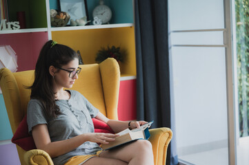Portrait of young smart and nerdy girl wearing spectacles relaxing on couch at home reading a book...