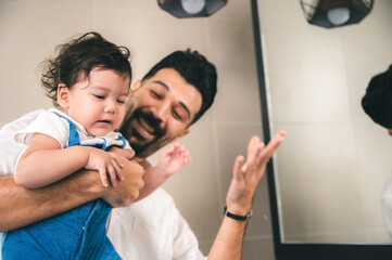 Handsome young father lifting small little daughter while training her about cleanliness by helping her wash her hands under running tap water in bathroom basin