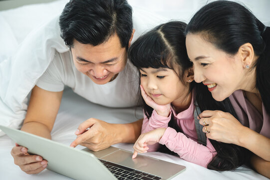 Cheerful young Asian family of handsome father and beautiful mother lying on bed with cute little daughter laughing while watching a comedy movie on laptop and relaxing at home