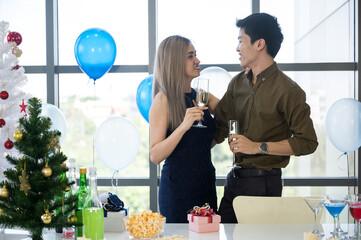 Closeup of hands of handsome man with beautiful woman celebrating and enjoying occasion with champagne and raising a toast while hugging and looking at camera at a decorated room with balloons