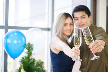 Closeup of hands of handsome man with beautiful woman celebrating and enjoying occasion with champagne and raising a toast while hugging and looking at camera at a decorated room with balloons
