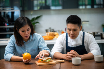 Cheerful and romantic young husband wearing apron drinking coffee with beautiful wife with sandwich and fruits at kitchen table while looking at each other