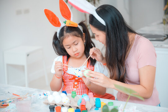 Beautiful young Asian woman wearing rabbit hairband painting eggs using paint colors in a creative work to celebrate easter with cute little daughter at home