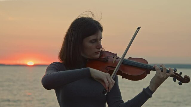 Young talented lady is playing violin skillfully, standing on rivershore and getting inspiration from pink sunrise, doing what she really loves, Foreground, Slow motion.