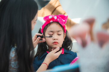 Young beautiful mother applying makeup and eye mascara with a brush while preparing innocent child for school program