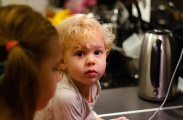 a small curly-haired blond boy looks seriously at the camera against the background of a kitchen table and a shiny kettle