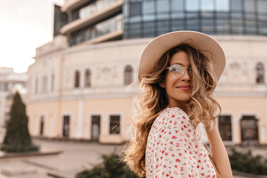  Gentle image sophisticated cute caucasian young lady on blurred light background of building. Blonde with long hair looks to side with slight grin in transparent glasses, beige hat and white blouse.