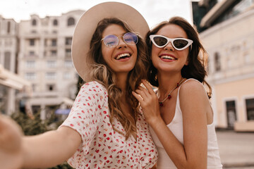 Happy caucasian young girls stand on street and take selfie using mobile phone. Blonde and brunette in glasses are dressed in light clothes. Loyal friends have wonderful moments of happiness and joy.