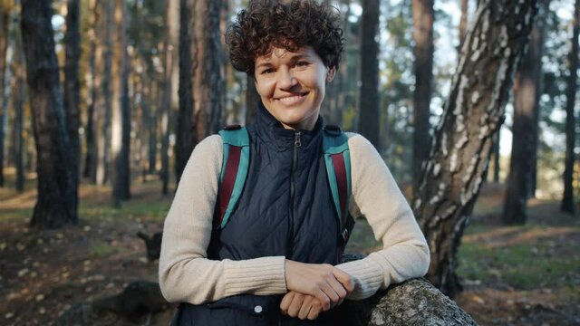 Slow motion portrait of female hiker with backpack smiling looking at camera standing in forest in autumn. People and travelling concept.
