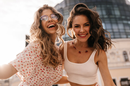 Close-up two graceful caucasian young girlfriends are smiling at camera embracing each other. Blonde in transparent glasses in blouse and brunette with red necklace in white top on street.