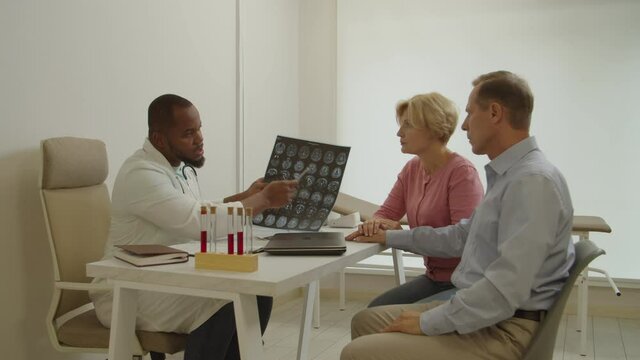 Professional black man doctor holding mri brain scan, explaining test results and treatment to worried middle aged male patient, supported by loving wife while mature couple visiting medical clinic.