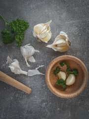 Bulbs of garlic (allium), fresh parsley and wooden kitchen mortar on concrete background. Overhead shot. Copy space.