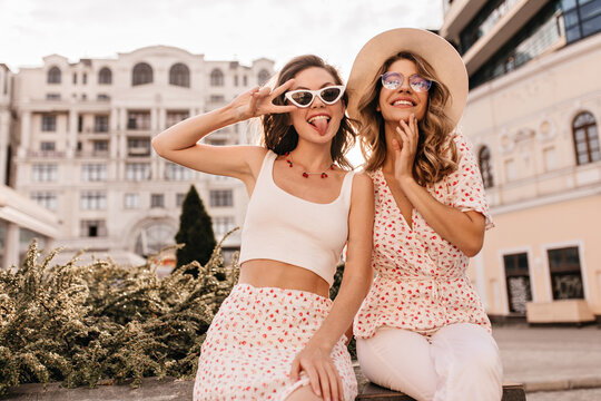 Funny caucasian young girls sit outdoors looking at camera. Brunette shows tongue and peace sign near her face. Blonde woman in beige hat smiling with teeth. Girls are dressed in white casual clothes.