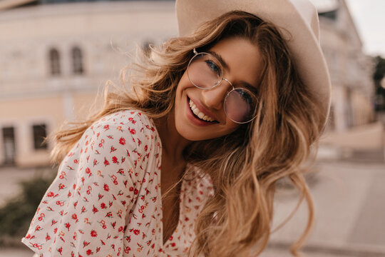 Close-up caucasian girl with snow-white smile looks into camera bending forward. Against blurred background of city, long-haired beauty in transparent round glasses, hat and blouse.