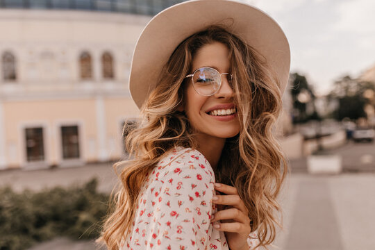 Close-up image of beaming caucasian young lady with flawless smile outdoors. Magic blonde with long hair in transparent glasses and beige hat. Travel, vacation and rest concept.