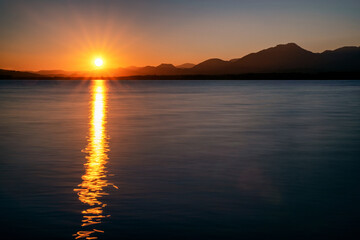 Reflecton of sunrays on water surface. Sun and silhouette of mountains at background