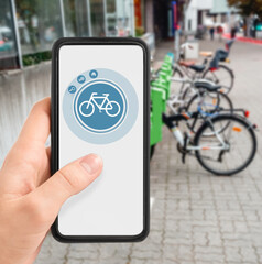 technology, transport and sustainability concept - close up of hand holding smartphone with mobile app and bicycle icon on screen over electric bike parking and charging station in city on background