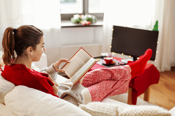 christmas, winter holidays and leisure concept - young woman reading book and resting her feet on table at cozy home