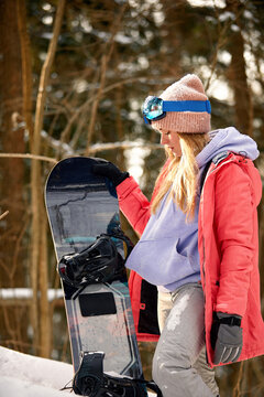 Caucasian woman snowboarder holds snowboard on beautiful snowy forest background in sunny day.
