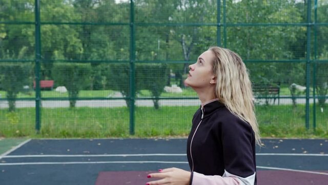 Beautiful girl throws ball into hoop on fenced sports ground. Blonde playing basketball or volleyball on court. Basketballer, hoopster, player trying to hit into rim. Sportswoman practices drills