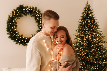 A happy man and a woman in love in sweaters embrace, relax and drink coffee with marshmallows sitting on the sofa in decorated room with a Christmas tree on a holiday in a cozy house. Selective focus