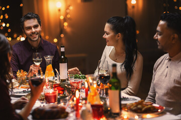 holidays, party and celebration concept - multiethnic group of happy friends having christmas dinner at home and drinking wine
