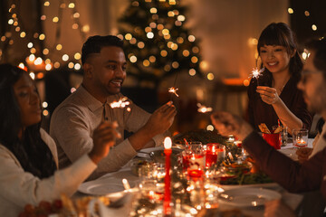 holidays and celebration concept - multiethnic group of happy friends with sparklers having christmas dinner at home