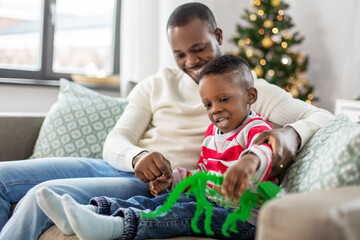 Fototapeta premium family, winter holidays and people concept - happy smiling african american father and baby son with dinosaur toy at home on christmas