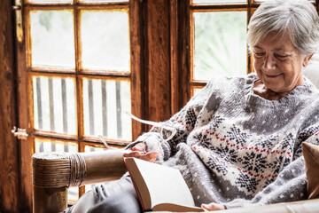 Portrait of senior woman sitting at home on armchair reading a book wearing a warm sweater and...
