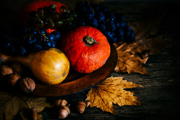 Naklejka na ściany i meble Thanksgiving composition with autumn fruit in wooden plate with maple leaves. Pumpkin and grapes vintage still life scene. November festive holiday food decoration. Thanksgiving dinner invitation card