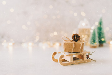 Eco Natural Christmas gift on toy wooden sled on white background with glowing light bokeh, copy space. New year card. Xmas decoration with house, Christmas tree and snow