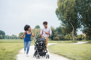 Happy black family having fun running with stroller outdoor at city park - Focus on faces