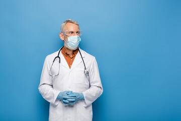 middle aged doctor in white coat, medical mask and goggles standing with clenched hands on blue