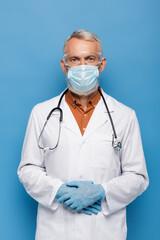 middle aged doctor in white coat, medical mask and goggles standing on blue