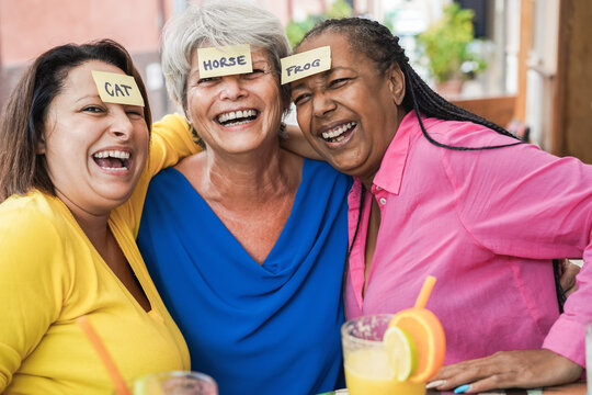 Portrait of multiracial senior women having fun playing guessing forehead game at bar - Focus on sticky notes on head