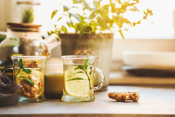 Two glass cups with fresh lemon and ginger tea on kitchen table with ginger root, herbs and kitchen...