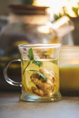 Glass cup of fresh brewed ginger tea with mint leaves and lemon on kitchen table at window...