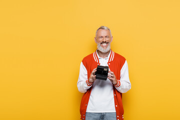 joyful middle aged man in bomber jacket holding black vintage camera isolated on yellow