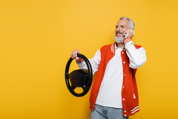 happy middle aged man holding steering wheel while imitating driving and talking on smartphone isolated on yellow