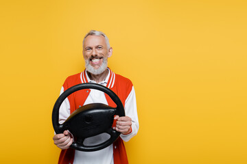 joyful middle aged man in bomber jacket holding steering wheel while imitating driving isolated on...