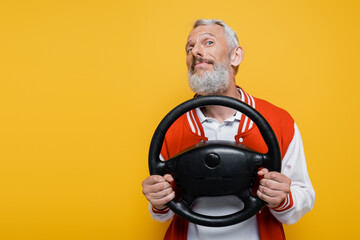 pleased middle aged man in bomber jacket holding steering wheel while imitating driving isolated on yellow