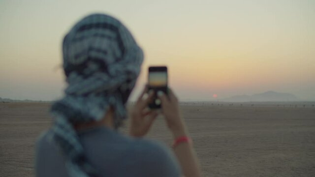 Woman in checkered keffiyeh taking picture of sunrise on mobile phone standing in the desert. Caucasian female tourist enjoy desert adventure.