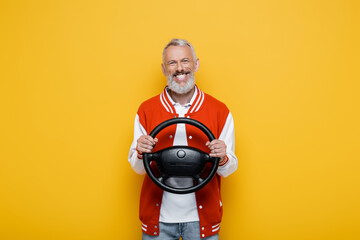happy middle aged man in bomber jacket holding steering wheel isolated on yellow - obrazy, fototapety, plakaty