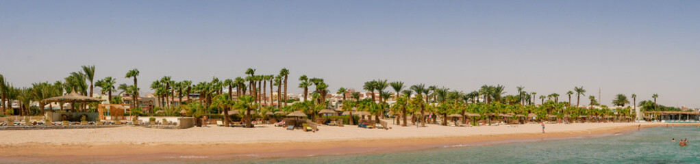Hurghada, Egypt - September 22, 2021: Panoramic view of the sandy Egyptian beach with green palms. People relax, sunbathe on sun loungers and swim in the Red Sea.