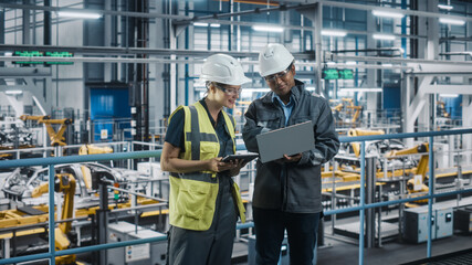 Male Specialist and Female Car Factory Engineer Talking and Working on Laptop and Tablet Computers. Automotive Industry 4.0 Facility Working on Vehicle Production. Diversity on Assembly Plant.