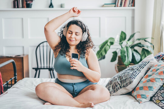 Overweight young woman with loose hair in top and shorts listens to music with wireless headphones holding smartphone in hand and dances sitting on double bed